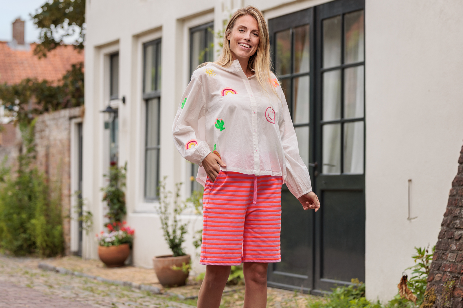 Persoon in een witte blouse met kleurrijke accenten en een roze gestreepte korte broek staat glimlachend voor een woning.