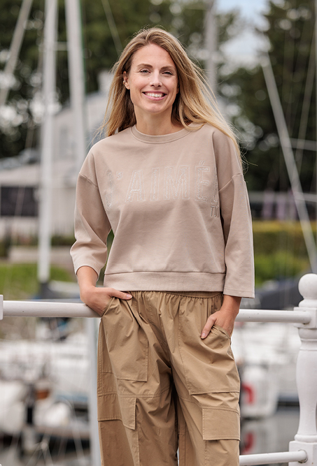 Een glimlachende vrouw met lang blond haar staat buiten bij een jachthaven. Ze draagt een beige trui met driekwartmouwen en een lichtbruine cargobroek. Op de achtergrond zijn onscherpe zeilboten, masten en groen te zien.