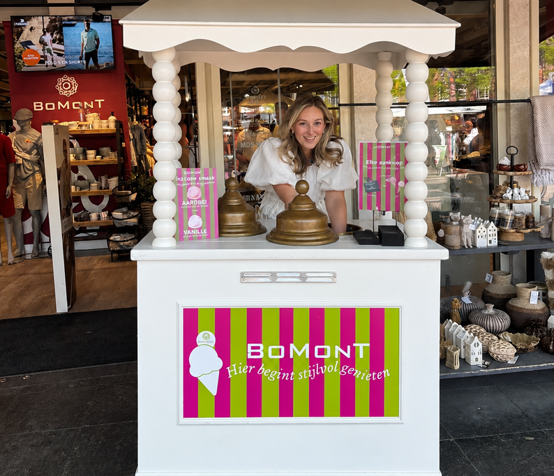 Vrouw staat glimlachend achter een witte ijskar met het logo van Bomont in een kleding- en woonwinkel.