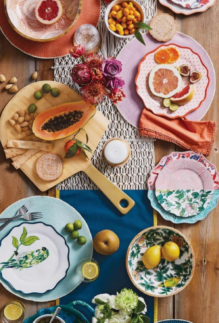 Bovenaanzicht van een kleurrijk gedekte houten tafel met verschillende borden, schalen en glazen. Op tafel liggen onder meer een gehalveerde papaja, citrusvruchten, brood, olijven en bloemen, gecombineerd met kleurrijk servies en textiel.