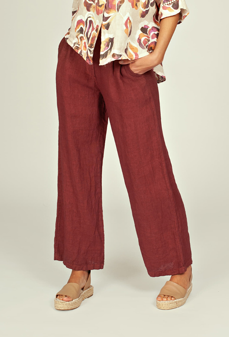 Persoon draagt een wijde linnen broek in een diepe roodbruine kleur. De broek heeft rechte pijpen, een losse pasvorm en wordt gecombineerd met sandalen en een blouse met bloemenprint.