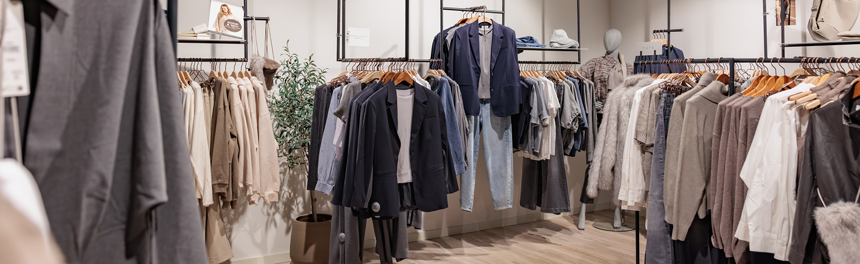 Kledingwinkel met rekken vol neutraal gekleurde kleding, zoals jassen, overhemden, broeken en truien, netjes gepresenteerd in een lichte, moderne winkelruimte.