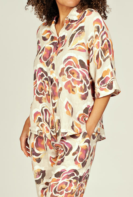Persoon draagt een luchtige linnen blouse met korte mouwen en een kleurrijke bloemenprint in tinten roze, beige en bruin. De blouse heeft een losse pasvorm en een V-hals met kraag, gecombineerd met een bijpassende broek