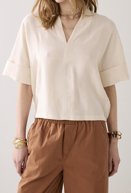 Persoon draagt een losvallende crèmekleurige blouse met korte mouwen en een V-hals, gecombineerd met een lichtbruine broek met elastische taille. De outfit wordt aangevuld met gouden armbanden en een fijne ketting.