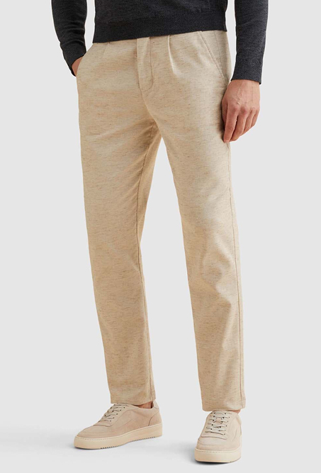 Persoon draagt een lichtbeige linnen broek met rechte pijpen, gecombineerd met een donker bovenstuk en lichtbeige sneakers. Achtergrond is lichtgrijs.