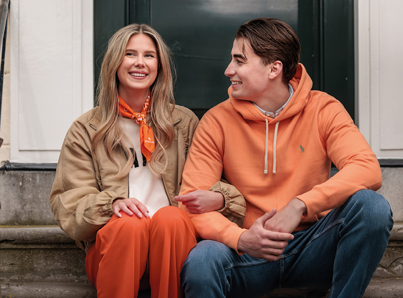 Twee jongeren zitten naast elkaar op een stenen trap voor een donkere deur. De vrouw draagt een beige jas, oranje broek en een oranje sjaaltje. De man draagt een oranje hoodie en een spijkerbroek. Ze glimlachen en kijken naar elkaar.