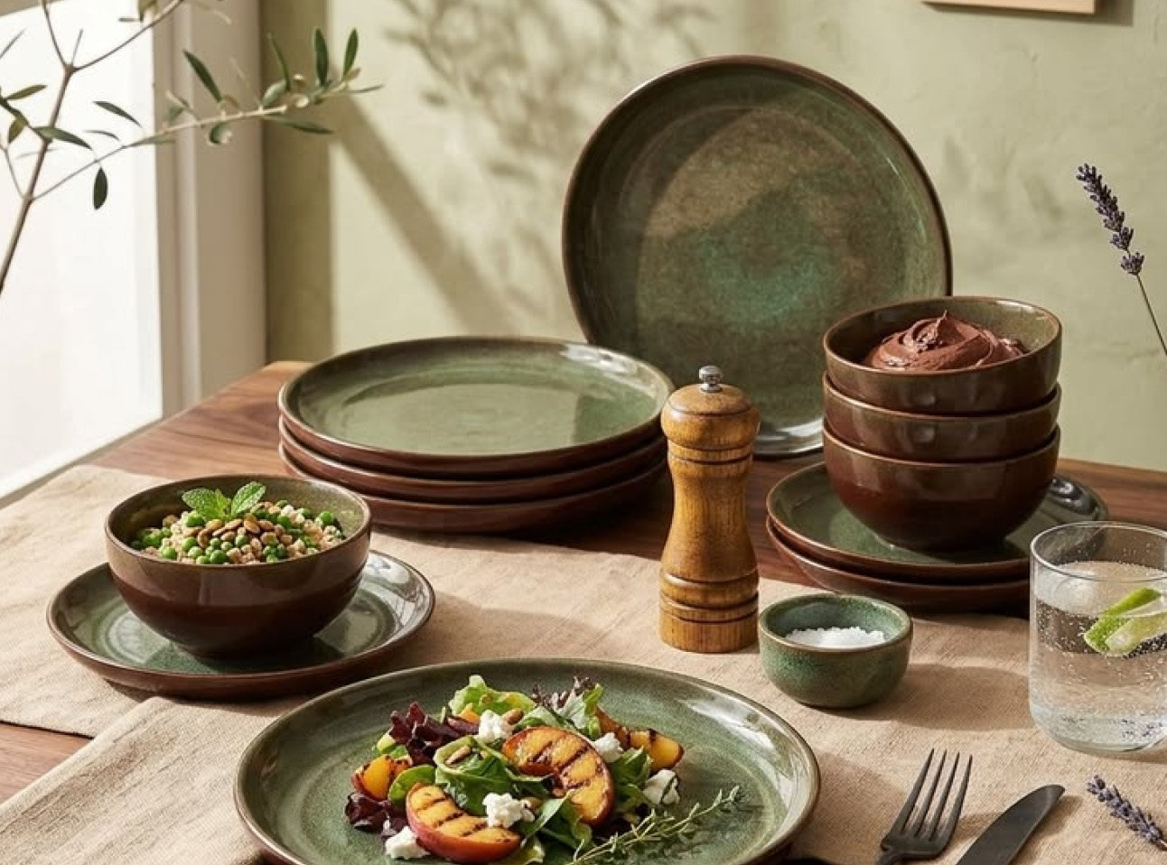 Gedekte houten eettafel met groen-bruin keramisch servies, een houten pepermolen en glazen. Op de voorgrond staat een bord met een kleurrijke salade en gegrilde perzik. Zacht zonlicht en natuurlijke tinten geven de setting een warme uitstraling.