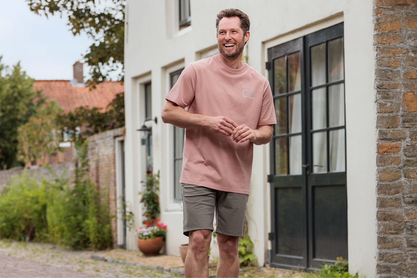 Persoon in een oudroze T-shirt en grijze korte broek loopt glimlachend over een pad langs een woning.