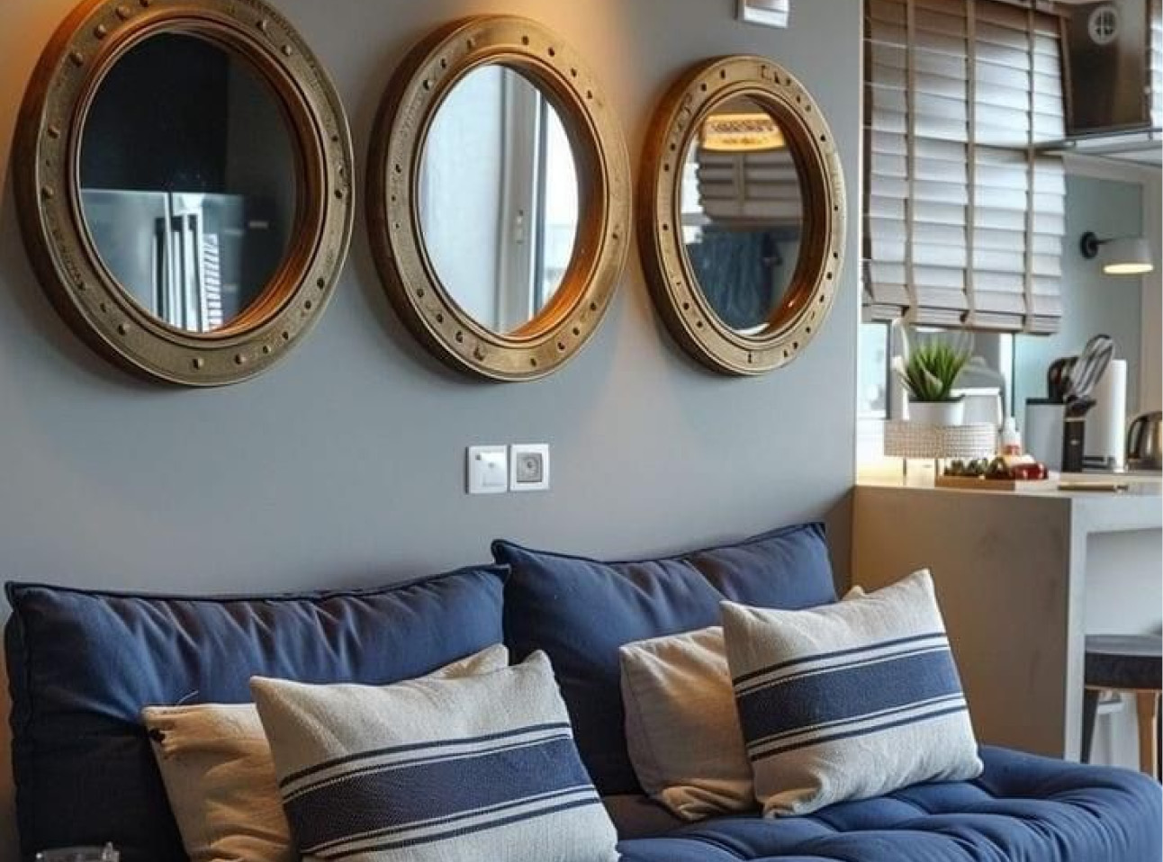 Woonkamer met een donkerblauwe bank en beige-blauw gestreepte sierkussens. Boven de bank hangen drie ronde spiegels met brede, bronskleurige lijsten. Rechts is een lichte keukenhoek met houten jaloezieën en een kleine plant zichtbaar.